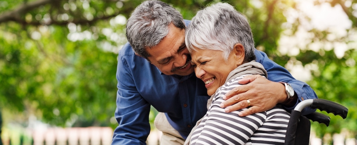 What Are Mass Tort Damages and Their Impact? A senior couple hug and laugh, with the woman being in a wheelchair, in a park by trees.