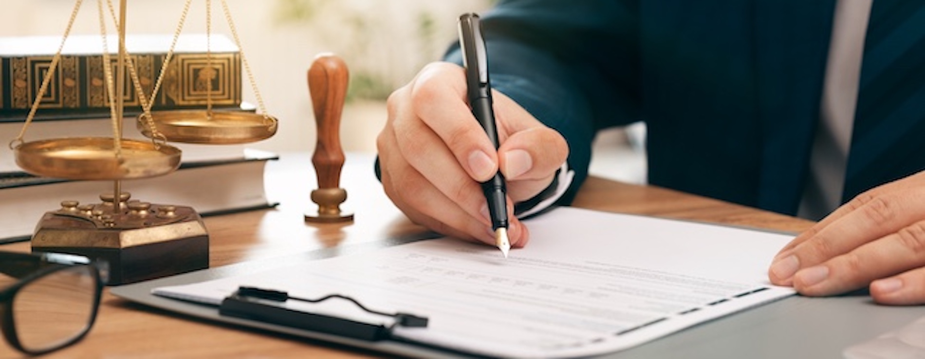 Choosing the Right Mass Tort Lawyer for Your Case Caucasian male lawyer signing paperwork in his office at his desk. Glasses and a scale are also on the desk.