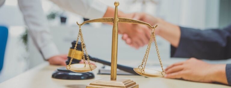 Two Caucasian people shaking hands over a desk. The gavel and scale are sitting on top of the desk.