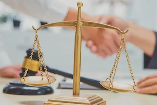 Two Caucasian people shaking hands over a desk. The gavel and scale are sitting on top of the desk.