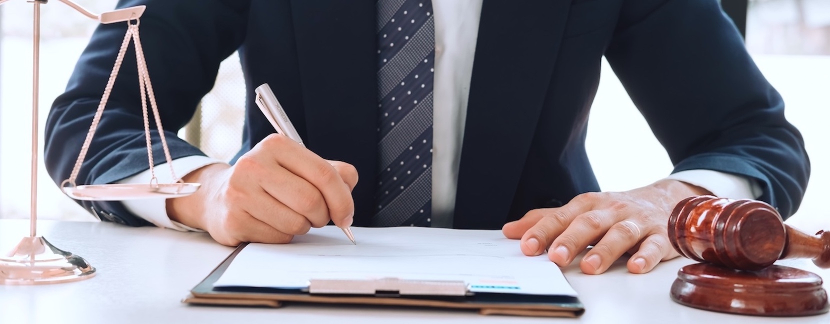 Pharmaceutical Negligence Claims: What You Need to Know A Caucasian male lawyer signing paperwork at his desk that has a scale to the left and a gavel to the right.