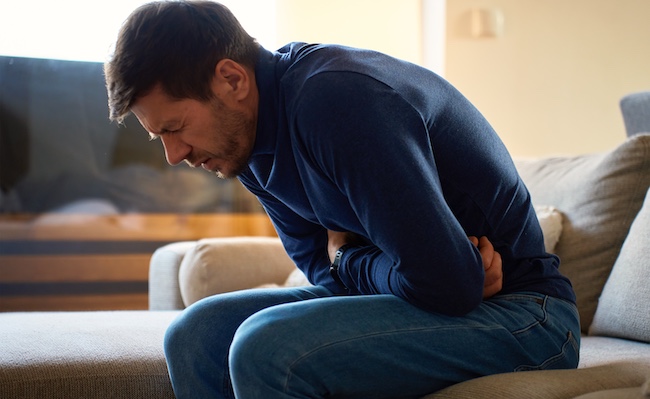 A man is experiencing severe abdominal pain on the couch, representing a victim of defective medical cases involving harmful drugs or devices.