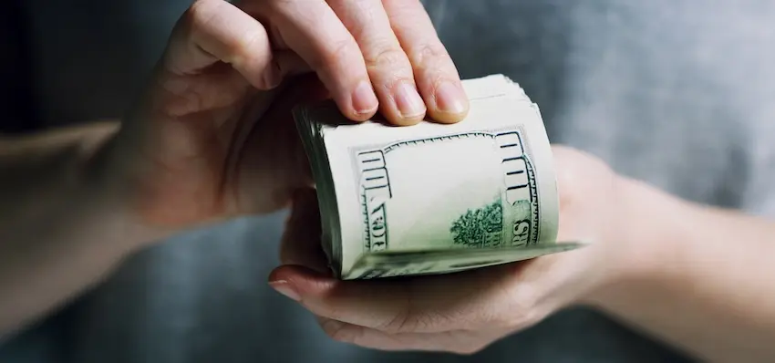 One hand passes a thick roll of hundred-dollar bills into another open palm against a blurred gray background representing the exchange of money in a first settlement offer that may fall far short of what an accident victim is truly owed for their injuries and losses in California.