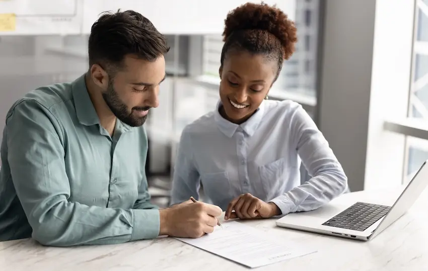 A bearded man in a teal shirt signs a document at a white marble countertop while a smiling woman in a white button-down shirt leans in beside him with a laptop open nearby illustrating the moment an accident victim reaches a satisfactory agreement and signs off on a negotiated settlement after rejecting the first settlement offer with guidance from Rueb Stoller Daniel.