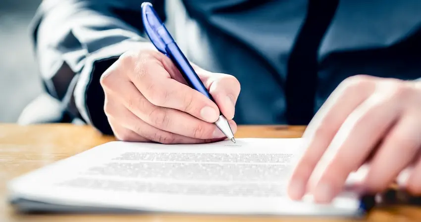 A close-up of a hand gripping a blue pen and signing a printed legal document on a wooden desk with another hand steadying the page representing the critical moment of signing paperwork related to an accident claim and why victims should always consult an attorney before accepting the first settlement offer from an insurer in California.